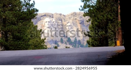 Stunning framed view of Mount Rushmore National Memorial, the iconic granite mountain carved with the faces of four U.S. presidents – George Washington, Thomas Jefferson, Theodore Roosevelt, and Abraham Lincoln. The monumental sculpture rises majestically, framed by tall lush pine trees and seen from a scenic paved road in South Dakota’s Black Hills. Soft sunlight highlights the stone details, blending American heritage, monumental artistry, and natural forest beauty.