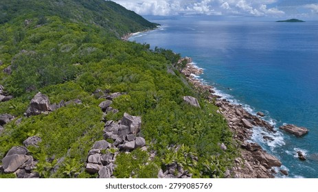 Stunning high-angle drone shot capturing the wild, forested coastline and granite boulders meeting the deep blue ocean waves near Anse Cocos, La Digue, Seychelles.