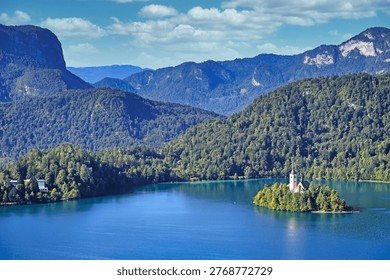 A stunning high-angle panoramic view of the emerald green Lake Bled featuring the iconic Pilgrimage Church of the Assumption of Maria on a small island, surrounded by the lush green Julian Alps