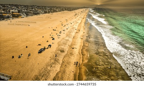 A stunning high-angle perspective of a bustling sandy beach during a golden sunset. Gentle turquoise waves roll onto the shore while people relax, walk, and play near coastal homes and volleyball courts under a warm, hazy sky in California.