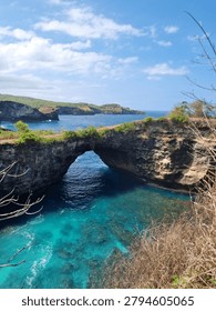 Stunning high-angle view of Broken Beach (Pasih Uug) in Nusa Penida, Bali, Indonesia. The image features the famous natural limestone archway over the vibrant, crystal-clear turquoise ocean water. Coral reefs are visible beneath the pristine sea, while dramatic coastal cliffs covered in green shrubbery stretch into the distant blue horizon under a bright, partly cloudy sky. This iconic tropical landmark perfectly showcases the exotic volcanic topography and marine beauty of the Indonesian islands.