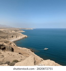 a stunning high-angle view of dramatic desert cliffs curving alongside deep, vivid blue waters under a clear sky. A small white boat glides across the serene sea near the rugged, rocky coastline, adding a touch of adventure to the vast landscape