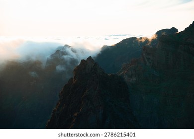 A stunning high-angle view of a prominent, sharp mountain peak standing tall against a dramatic landscape of deep canyons and volcanic ridges. Light mist and a dense sea of clouds roll over the distant peaks, beautifully catching the warm, golden glow of the setting sun. This atmospheric and majestic scene perfectly captures the untamed spirit of high-altitude exploration and nature's raw beauty, making it ideal for travel, adventure lifestyle, and wilderness themes.