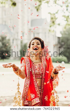 Stunning Indian bride dressed in Hindu traditional wedding clothes lehenga embroidered with gold and a veil smiles tender posing outside with golden accessories under the rain of petals