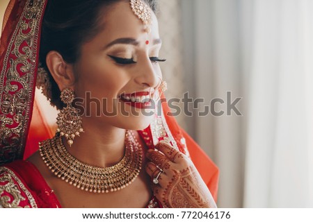 Stunning Indian bride dressed in Hindu red traditional wedding clothes lehenga embroidered with gold and a veil smiles tender