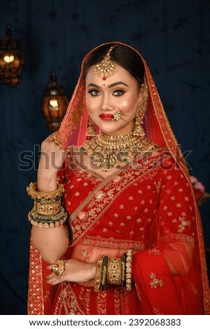 Stunning Indian bride dressed in traditional red bridal lehenga with heavy gold jewellery and veil smiles tenderly in studio lighting. Wedding fashion and lifestyle.