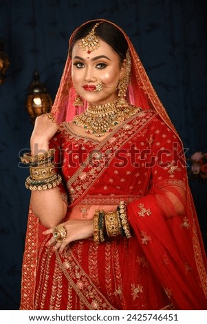 Stunning Indian bride dressed in traditional red bridal lehenga with heavy gold jewellery and veil smiles tenderly in studio lighting. Wedding fashion and lifestyle.