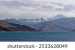 A stunning landscape of a mountain range with snow-capped peaks and a serene blue lake under a partly cloudy sky.