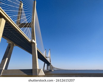 Stunning lowangle view of a modern cablestayed bridge stretching across a calm body of water under a vibrant blue sky.  Ideal for architecture, travel, engineering, and infrastructure projects.