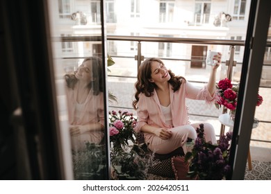 Stunning madame enjoying her morning drink, sitting nearby peonies bouquets. Brunette lady wearing lovely pink homewear and new raspberry lipstick posing for camera on the open balcony