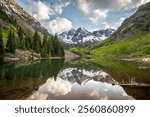 The stunning Maroon Bells mountains rise above a tranquil lake, surrounded by lush greenery and snow-capped peaks under a bright blue sky.