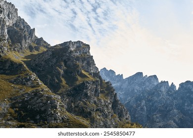 A stunning mountain landscape featuring rugged cliffs and rocky peaks under a blue sky with wispy clouds. The foreground showcases lush greenery and rocky terrain, creating a dramatic natural scene. - Powered by Shutterstock