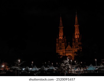 A stunning nighttime view of a neo-Gothic cathedral with illuminated spires, casting a warm orange glow against the dark sky. The iconic twin towers are framed by trees and lights in the foreground. - Powered by Shutterstock