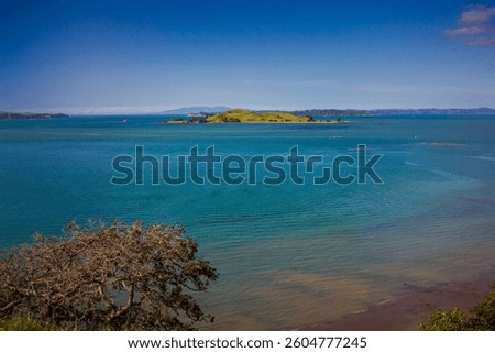 A stunning ocean view featuring a red ferry boat sailing near lush green islands in Auckland, New Zealand. The deep blue water contrasts beautifully with the vibrant landscape under a clear blue sky.