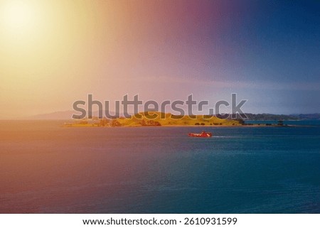 A stunning ocean view featuring a red ferry boat sailing near lush green islands in Auckland, New Zealand. The deep blue water contrasts beautifully with the vibrant landscape under a clear blue sky.