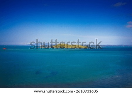 A stunning ocean view featuring a red ferry boat sailing near lush green islands in Auckland, New Zealand. The deep blue water contrasts beautifully with the vibrant landscape under a clear blue sky.