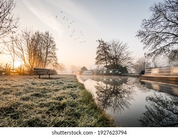 Stunning Old Canal House Boats Landscape Sunrise In Countryside With River And Single Lone Wooden Bench. Frost On Grass Reflection Of Trees And Vessels Chimney Smoke In Still Calm Water Birds In Sky