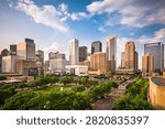 Stunning panoramic elevated view of the downtown Houston cityscape skyline featuring modern high-rise skyscrapers and commercial architectural towers under a vibrant blue sky with soft white dramatic clouds. The foreground highlights a lush green urban park filled with dense tree canopies, scenic city streets, and well-designed roads creating a striking contrast between nature and urban development. Shot during late afternoon daylight, the warm sunlight illuminates the glass and concrete facades of the corporate buildings in Texas, USA. Perfect visual for themes related to real estate, urban planning, American cities, business districts, financial hubs, metropolitan lifestyle, tourism, commercial growth, eco-friendly green cities, architectural design, corporate headquarter locations, travel destinations, and Texas economic development. High-quality urban landscape image ideal for marketing campaigns, editorial news articles, travel brochures, website banners, and corporate presentation backgrounds.