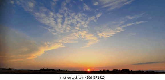 Stunning panoramic landscape of a dramatic sunset with a deep blue sky and scattered clouds illuminated by warm orange and golden light. A silhouette of a rural town or village is visible on the horiz
