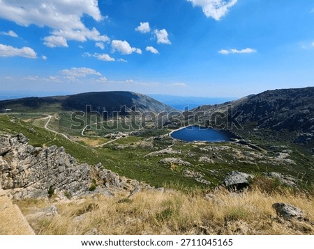 A stunning panoramic view of the Covão do Meio reservoir nestled in a lush green valley in the Serra da Estrela Natural Park, Portugal. A winding road snakes through the mountainous landscape under a bright blue sky with scattered clouds.