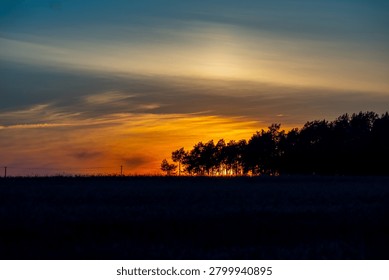 A stunning panoramic view of a dramatic sunset glowing over a dark field silhouette, with the sun dipping behind a row of trees under an expansive sky filled with shifting layers of gold, orange, and deep blue twilight tones.