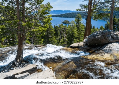 A stunning panoramic view from Eagle Falls overlook at Emerald Bay State Park, Lake Tahoe, California. Crystal-clear water rushes over smooth granite rocks in the foreground, creating a silky white cascade framed by towering pine trees with reddish-brown bark. In the middle distance, the deep cobalt-blue waters of Emerald Bay stretch toward the horizon, with the iconic Fannette Island — the only island in Lake Tahoe — rising from its center. Dense evergreen forests blanket the surrounding hillsides, and distant mountain ridges fade under a bright blue sky with wispy clouds. The layered composition combines dynamic flowing water, lush forest, serene alpine lake, and majestic mountain scenery, making this image ideal for travel, tourism, nature, outdoor recreation, and California destination campaigns.