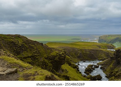 A stunning panoramic view of an Icelandic canyon, showcasing moss-covered cliffs, a flowing river, and expansive green plains stretching towards the horizon under a cloudy sky. - Powered by Shutterstock