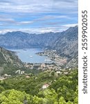 A stunning panoramic view of Kotor Bay, Montenegro, featuring calm blue waters, green hills, towering mountains, and a charming coastal town dotted with ships and winding roads.