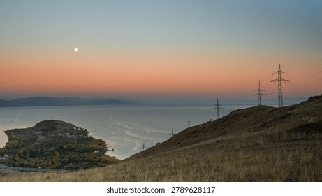 A stunning panoramic view of Lake Sevan during twilight, showing the glowing moon above the peaceful water and the silhouette of hills in soft pastel hues.