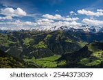 Stunning panoramic view from Stanserhorn Mountain in Switzerland, showcasing the vibrant green valleys and snow-capped peaks of the Alps. The clear blue sky and scattered clouds enhance the majestic