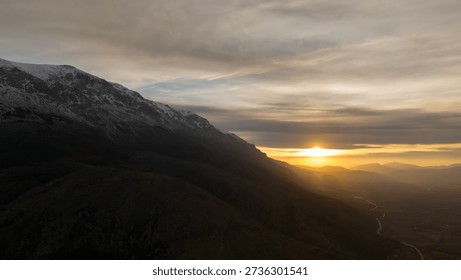Stunning panoramic view of a sunset behind a dark mountain ridge casting a golden glow over a vast valley under a dramatic cloudy sky in a remote natural landscape area