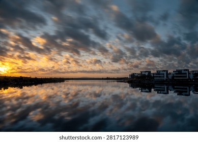 A stunning patchwork of altocumulus clouds ignites in gold and pink at sunset, perfectly mirrored in the glassy surface of an urban waterway flanked by modern residential buildings. Long exposure smooths the water surface, enhancing the symmetrical reflection of dramatic clouds and city silhouette. This image captures the surprising natural beauty of urban landscapes during golden hour.