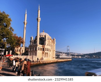 stunning photograph of a waterfront mosque with a bridge and cityscape in the background. The photo captures anonymous people walking on a public street during sunset, with no visible faces