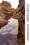 Stunning reflection of clouds and sky in a calm tide pool surrounded by dark volcanic shore rocks.Clear water puddle among coastal rock formations creating a beautiful natural mirror of the cloudy sky.Low angle vertical shot of sky reflection on a serene rocky beach tide pool during low tide.