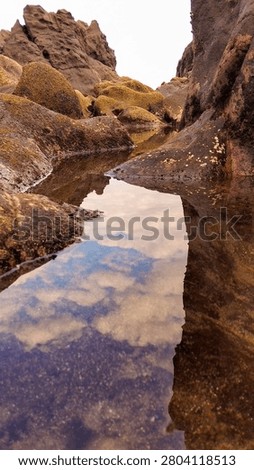 Stunning reflection of clouds and sky in a calm tide pool surrounded by dark volcanic shore rocks.Clear water puddle among coastal rock formations creating a beautiful natural mirror of the cloudy sky.Low angle vertical shot of sky reflection on a serene rocky beach tide pool during low tide.
