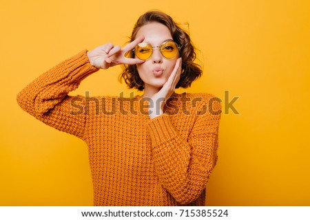 Stunning short-haired female model posing with kiss face expression on yellow background. Close-up portrait of stylish european girl standing with peace sign in front of wall.