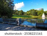 a stunning shot of a water fountain in the middle of silky green lake water surrounded by lush green and autumn colored trees with blue sky and clouds at Lenox Park in Brookhaven Georgia USA