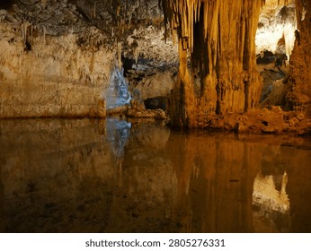 Stunning subterranean cavern interior featuring dramatic natural limestone formations. This professionally lit cave photograph showcases golden-hued stalactites hanging from the ceiling and corresponding stalagmite formations rising from the cave floor. A still underground pool creates perfect mirror reflections of the mineral deposits and cave walls, with warm amber and cream-colored mineral staining visible throughout. The cave passage extends into the distance with natural light visible at the far end. This natural wonder image is ideal for travel, geology, nature, adventure, and educational content. Perfect for tourism boards, geological publications, adventure travel websites, and nature documentaries.