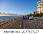 a stunning summer landscape along the beach at sunrise with blue and white lounge chairs, blue ocean water, palm trees, hotels, and footprints in the sand at Bal Harbour Beach in Miami Beach Florida