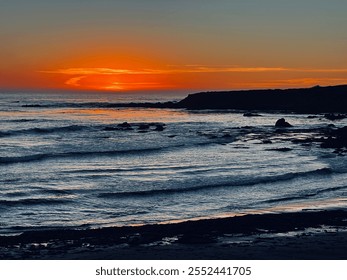 A stunning sunset casts a warm orange glow over a rocky shoreline, with gentle waves rolling in and a silhouette of a distant cliff. The scene exudes tranquility and natural beauty. - Powered by Shutterstock