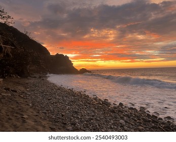 A stunning sunset over a beach with dramatic cloud. Gentle ocean waves break on the shore lined with stones and sand, while a dark rocky formation silhouette against the horizon.