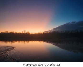 Stunning sunset over a calm river with vibrant colors reflecting on the water, creating a serene and tranquil atmosphere. Silhouetted trees along the horizon add depth to the peaceful twilight scene. - Powered by Shutterstock