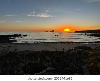 A stunning sunset over a rocky coastline, with the sun dipping below the horizon, casting an orange glow across the sky and reflecting on the calm ocean waters. - Powered by Shutterstock