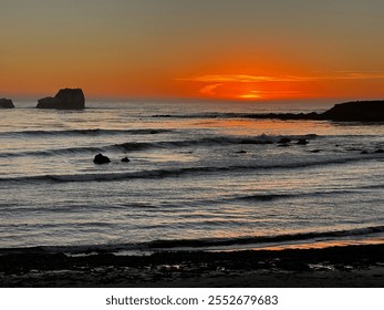 A stunning sunset over a rocky shoreline, with the sun casting an orange glow across the calm waves and silhouetted rock formations in the distance.  - Powered by Shutterstock