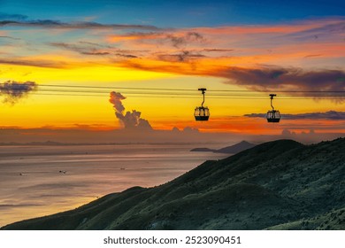 A stunning sunset view over the ocean with cable cars silhouetted against the vibrant sky. The sun is setting behind dramatic clouds, casting a warm golden glow across the water, with a distant bridge - Powered by Shutterstock