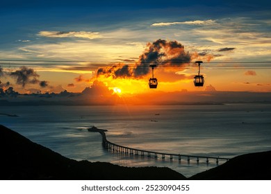 A stunning sunset view over the ocean with cable cars silhouetted against the vibrant sky. The sun is setting behind dramatic clouds, casting a warm golden glow across the water, with a distant bridge - Powered by Shutterstock