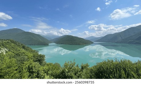 A stunning turquoise lake reflects green mountains and fluffy clouds, set beneath a clear blue sky, creating a peaceful, mirror-like scene - Powered by Shutterstock