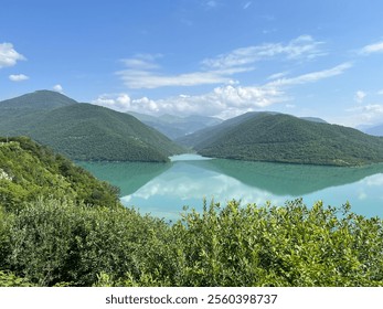 A stunning turquoise lake reflects green mountains and fluffy clouds, set beneath a clear blue sky, creating a peaceful, mirror-like scene - Powered by Shutterstock