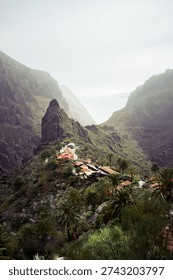 A stunning vertical composition of the iconic Masca village nestled within its deep volcanic ravine. This portrait orientation emphasizes the height of the surrounding cliffs and the steep drop-off of the valley. The hazy, soft light creates a dreamlike atmosphere, highlighting the contrast between the dark rocky textures and the vibrant green palm trees. The ocean is visible as a pale silhouette through the mountain pass.
