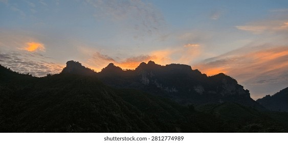 A stunning vertical view of the sun setting directly behind a dramatic, twin-peaked limestone mountain ridge in Na Hang, Tuyên Quang province, Vietnam. The brilliant golden glow contrasts sharply with the dark, silhouetted slopes in the foreground, creating a peaceful and majestic twilight atmosphere